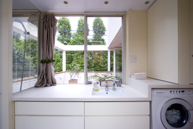 圧倒的に明るく、開放感がある洗面台。洗面台の正面には、テラス越しに緑の並木が広がる。鏡がないと不便だが、右側の壁（洗濯機上部）は扉になっていて内側に鏡が貼ってある。鏡を使わない時は景色を楽しみ、必要に応じて鏡も使えるアイデアだ
撮影：NewHouse出版