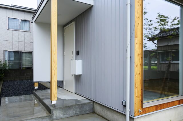 玄関ポーチ。柱を一番下まで落とすのではなく、袖壁や柱の下部に空きをつくり浮遊感を演出。住宅らしさを軽減した。画像右、木壁下部にある金具はアウターシェードを留めるもの。室内側からの視線の抜けは確保しながら日射を遮ることができる。日射遮蔽により冷房負荷の低減と冷房の効きやすさにもつながっている。