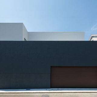 巨大な暗色の壁が印象的な正面外観。壁の奥に階段があり、玄関が道路から直接見えない。家の構成がわからないだけでなく、階段を登る時にワクワク感を感じられる空間になっている。周囲との調和を考えて黒ではない暗色とした配色は、重量感を与える意図もある