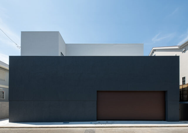 巨大な暗色の壁が印象的な正面外観。壁の奥に階段があり、玄関が道路から直接見えない。家の構成がわからないだけでなく、階段を登る時にワクワク感を感じられる空間になっている。周囲との調和を考えて黒ではない暗色とした配色は、重量感を与える意図もある
