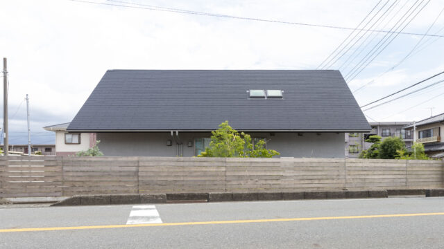 敷地の北を走る幹線道路から見た「サンカクヤネノイエ」。車の騒音や排気ガスから生活空間を守るために大屋根と木塀で覆い、1階の窓部分は植栽で目隠ししている。2階はほとんど窓がなく全体的に閉じた印象だが、邸内にはこの外観から想像もつかない明るい大空間が広がる