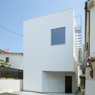 外壁には、白のガルバリウム鋼板（小波板）を採用。玄関ポーチは敷地背面の斜めの隣地境界ラインと平行に切り取り、シャープな造形美を強調させるモルタル仕上げ。窓の数をあえて絞ることでプライバシーを担保しつつも、室内への豊かな採光と視線の抜けをもたらしている。