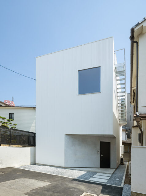 外壁には、白のガルバリウム鋼板（小波板）を採用。玄関ポーチは敷地背面の斜めの隣地境界ラインと平行に切り取り、シャープな造形美を強調させるモルタル仕上げ。窓の数をあえて絞ることでプライバシーを担保しつつも、室内への豊かな採光と視線の抜けをもたらしている。